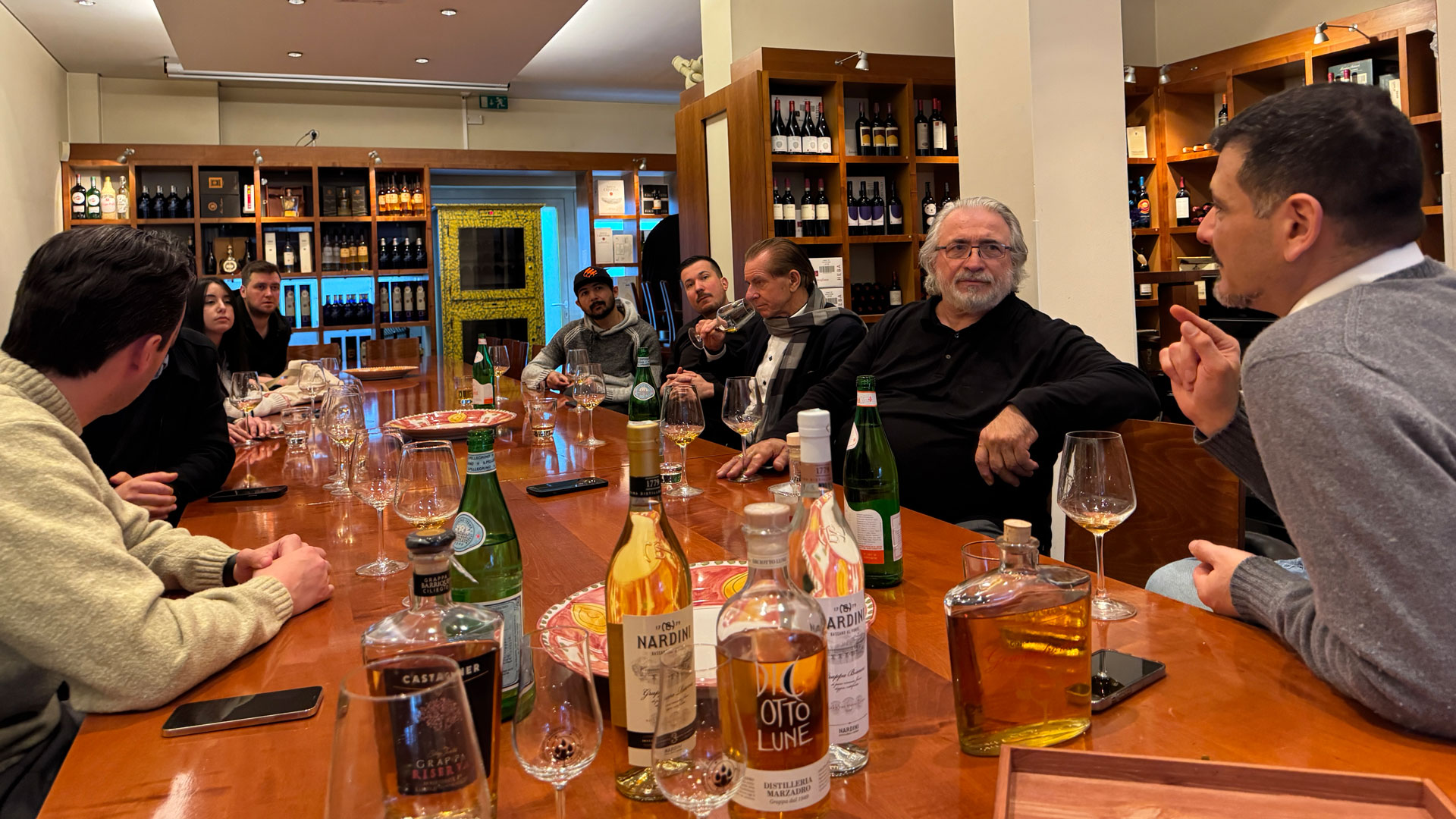 Foto: Grappaverkostung in geselliger Runde an einem langen Holztisch in stilvollem Wein- und Spirituosengeschäft.