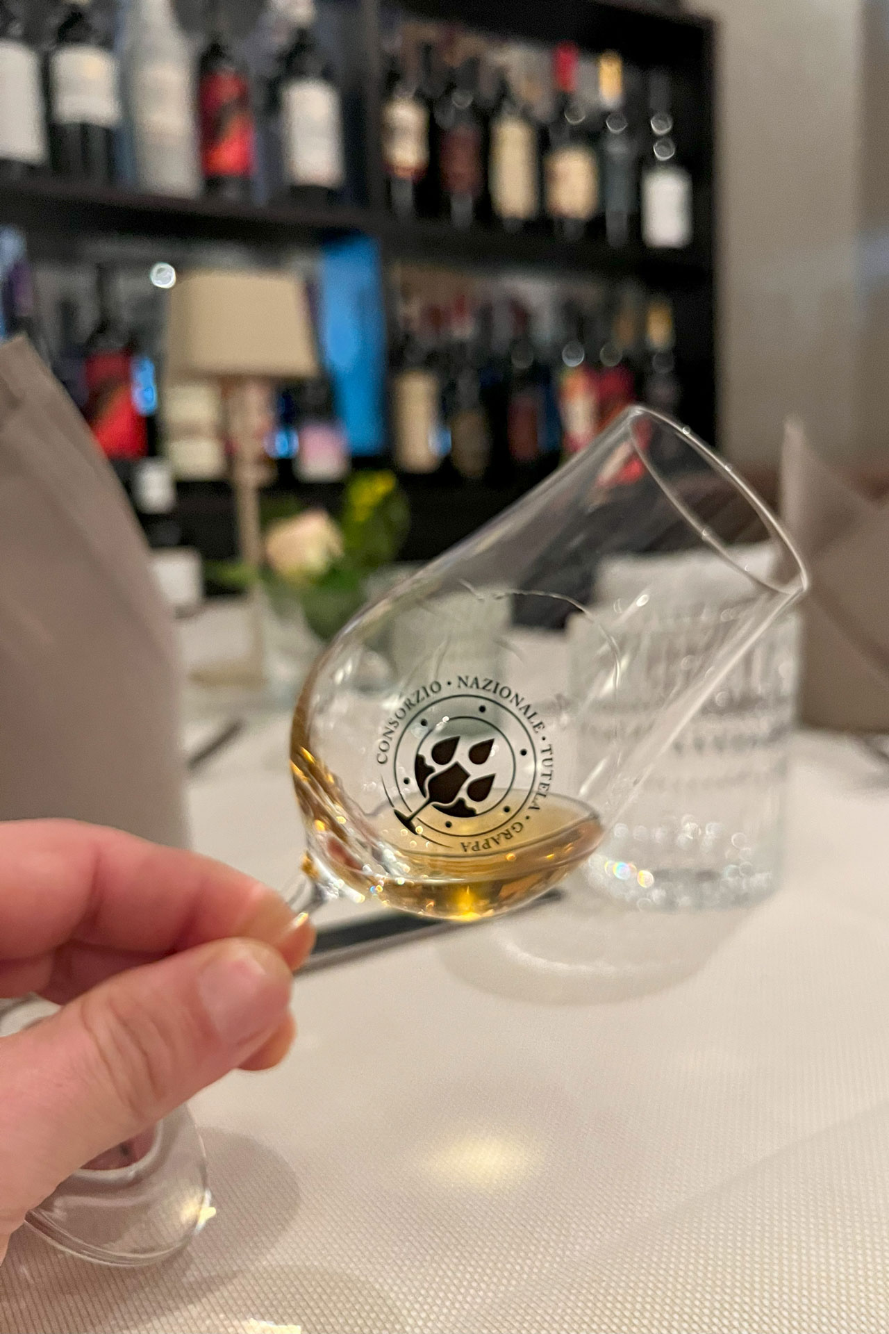 Foto: Hand hält Grappaglas mit goldfarbenem Inhalt und Logo des Consorzio Nazionale Grappa, im Hintergrund Weinregal und gedeckter Tisch.