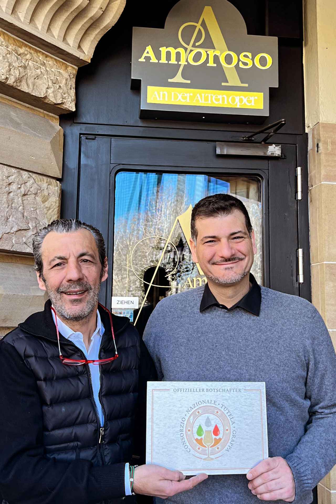 Foto: Zwei Männer vor dem Eingang des Restaurants „Amoroso“ mit Schild des Consorzio Nazionale Grappa als offizieller Botschafter in den Händen.