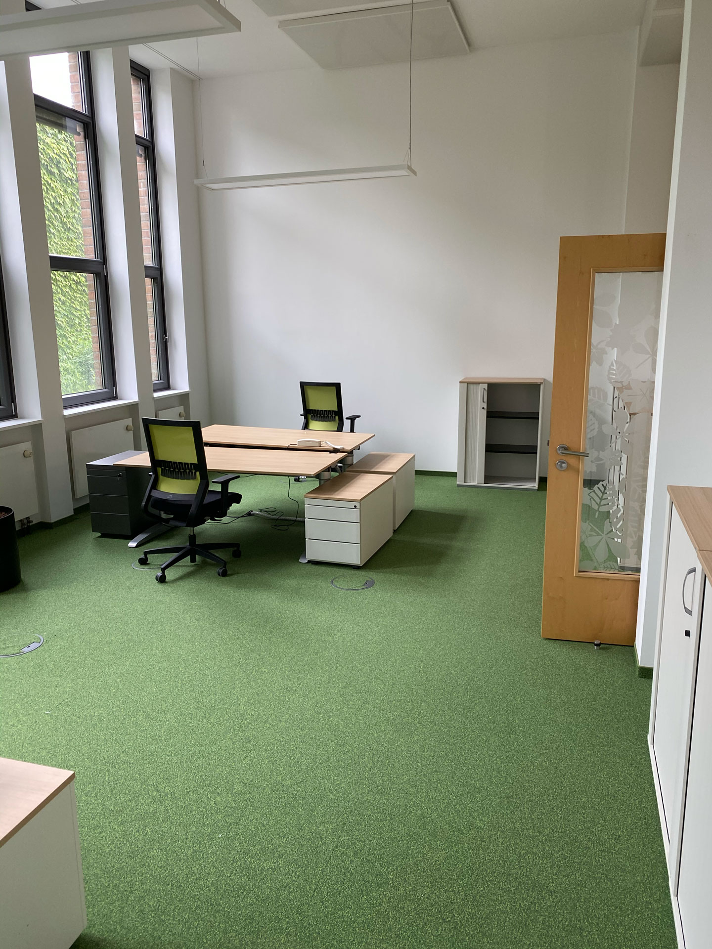 Foto: Modernes, helles Büro mit zwei Schreibtischen, grüner Teppichboden, großen Fenstern und minimalistischer Einrichtung.