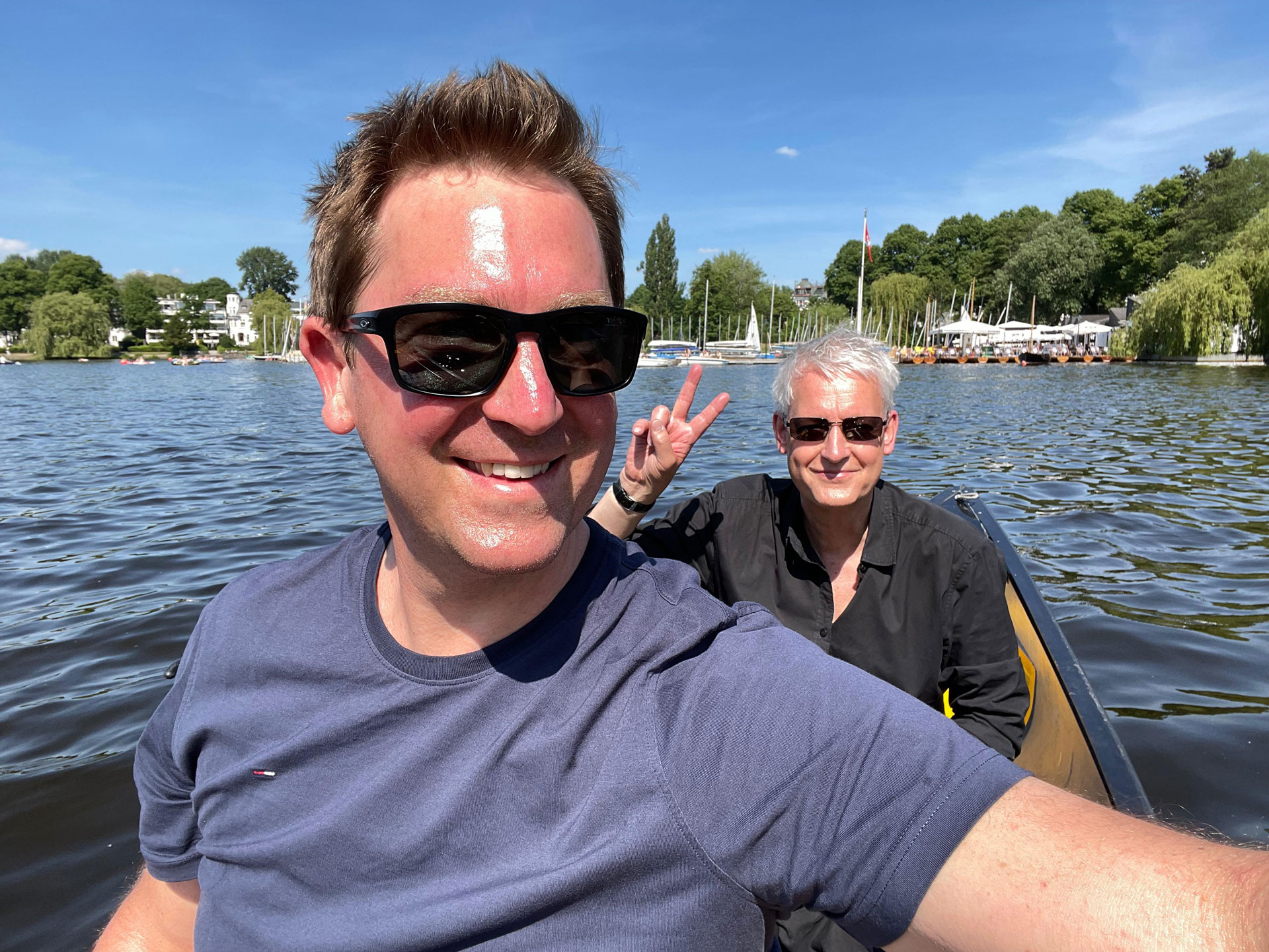 Foto: Zwei Männer sitzen lachend in einem Boot auf einem See an einem sonnigen Tag; der Vordergrund zeigt einen Mann mit Sonnencreme auf der Stirn, der andere macht ein Peace-Zeichen im Hintergrund.