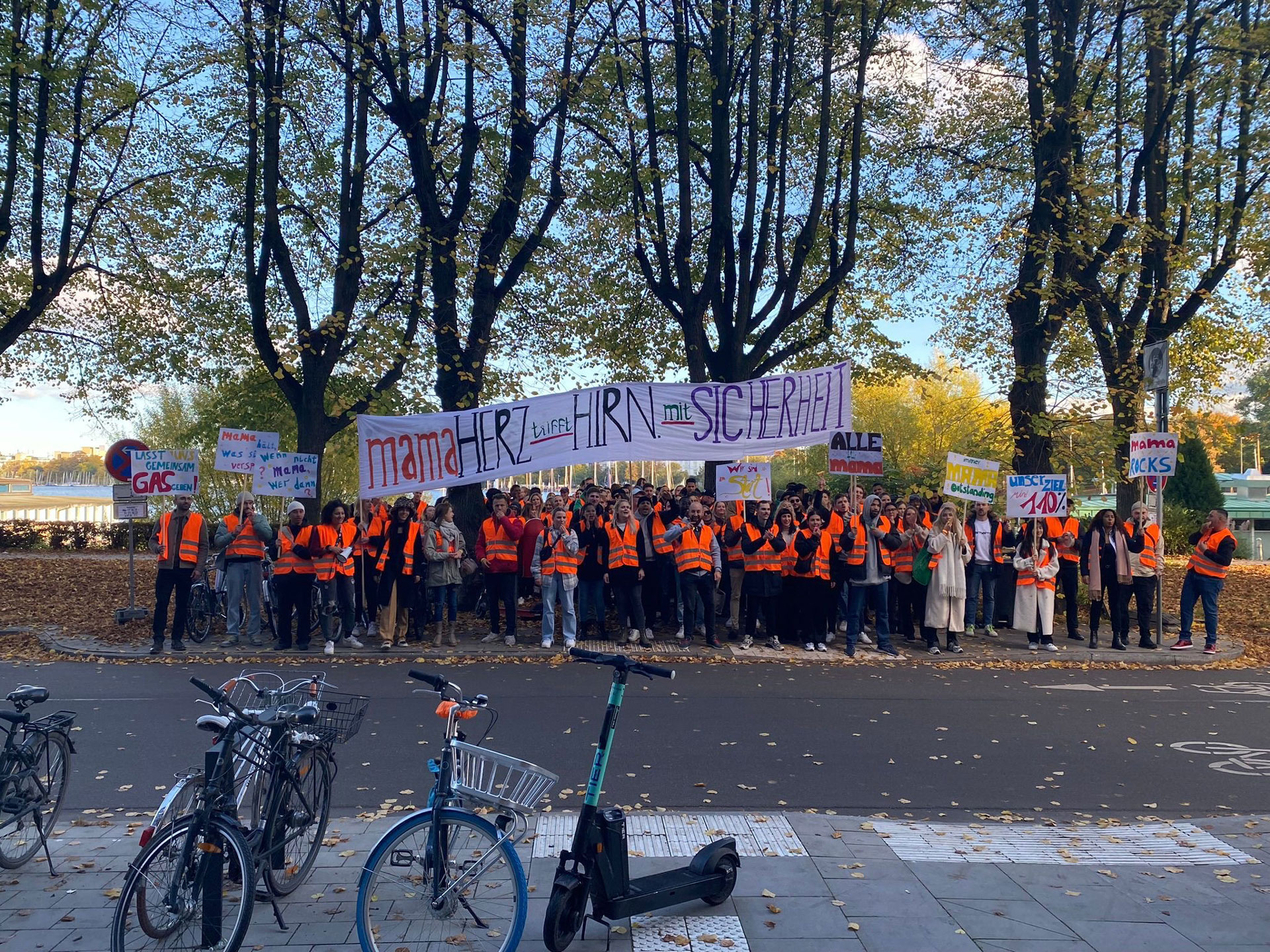 Foto: Große Gruppe von Menschen mit orangenen Warnwesten demonstriert friedlich auf einer Allee mit Plakaten wie „mamaHERZ trifft HIRN mit SICHERHEIT“ und „mama ROCKS“.