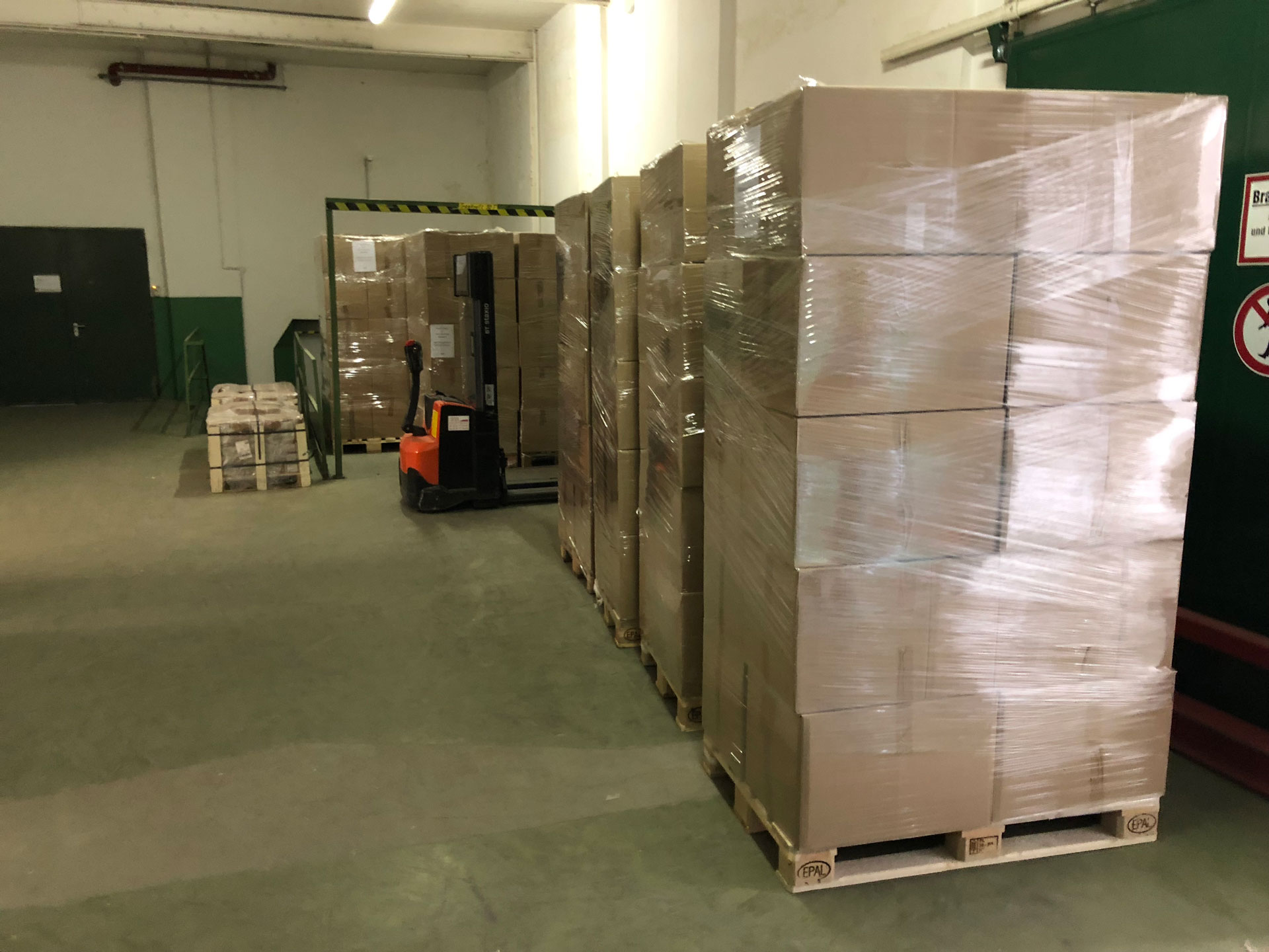 Foto: Lagerraum mit mehreren gestapelten Paletten voll verpackter Kartons, teilweise in Folie eingeschweißt, daneben ein Elektrohubwagen.