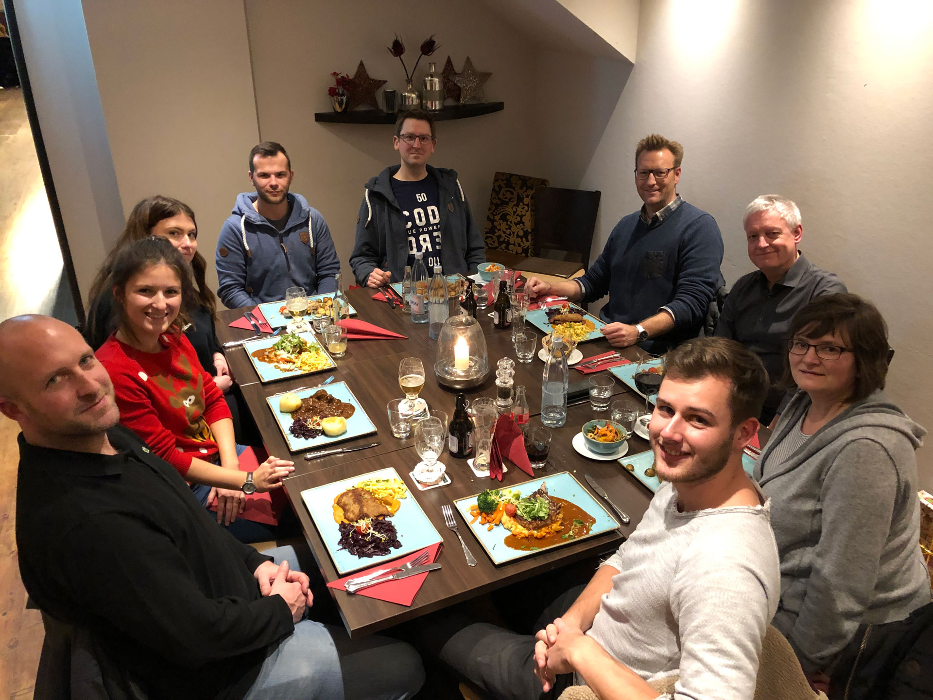 Foto: Zehn Personen sitzen fröhlich um einen festlich gedeckten Restauranttisch, genießen gemeinsames Essen und blicken gut gelaunt in die Kamera.