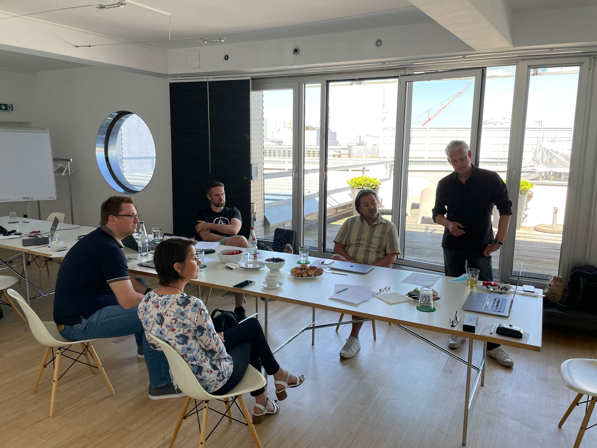 Foto: Fünf Personen sitzen und stehen in einem modernen, hellen Besprechungsraum mit Dachterrasse, bei einem Workshop oder Meeting mit Getränken und Snacks auf dem Tisch.