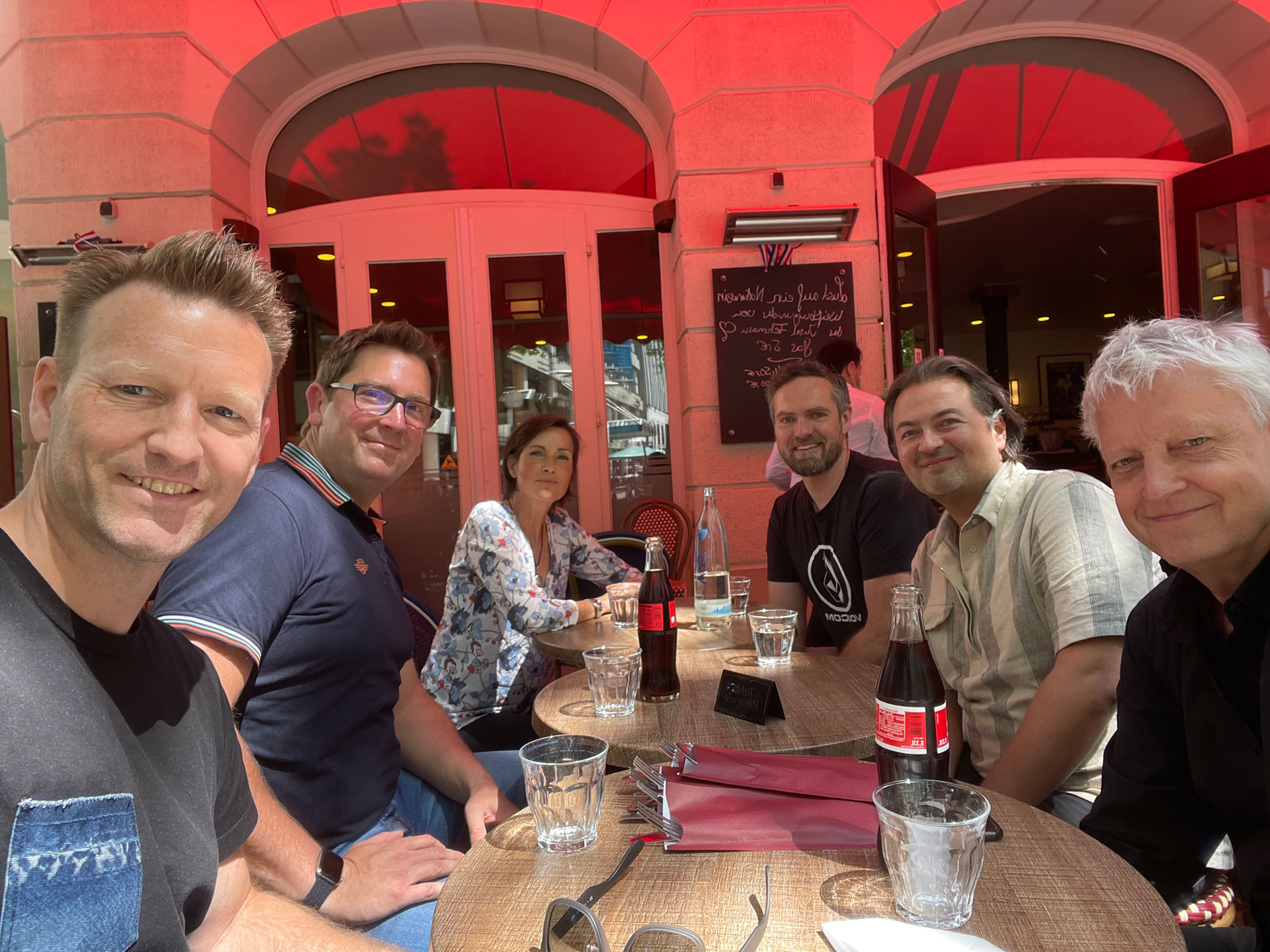 Foto: Sechs Personen sitzen gut gelaunt an einem runden Tisch im Außenbereich eines Cafés mit rotem Sonnenschutz, leere Gläser und Cola-Flaschen vor sich.