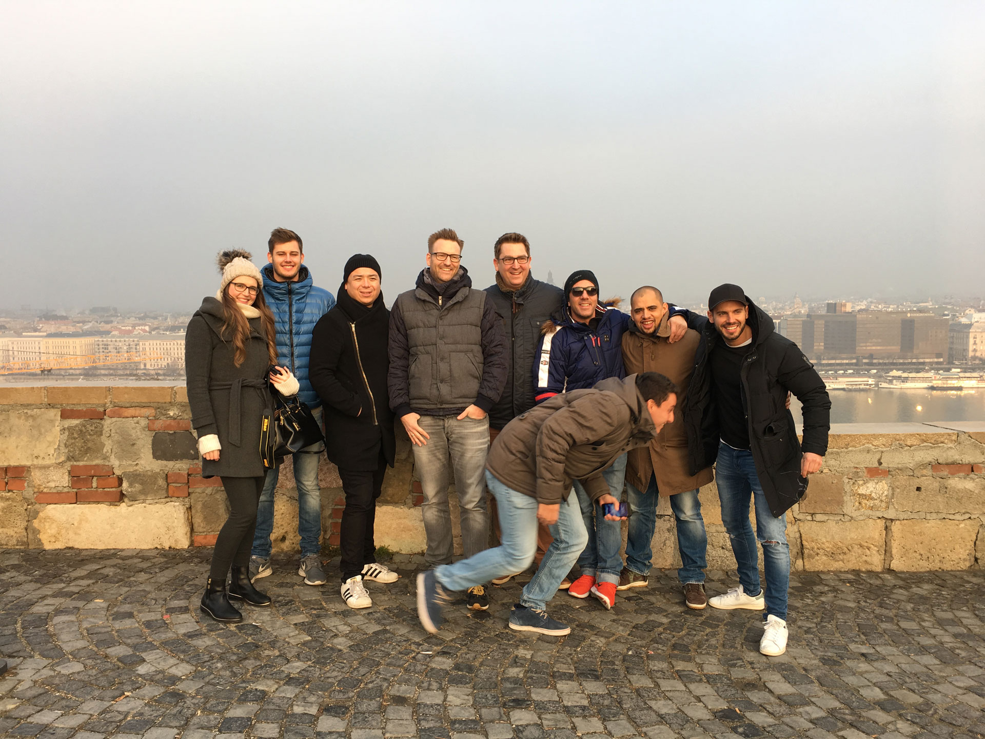 Foto: Neun Personen stehen lachend auf einem Aussichtspunkt mit Blick auf eine Stadt, eine Person läuft ins Bild.
