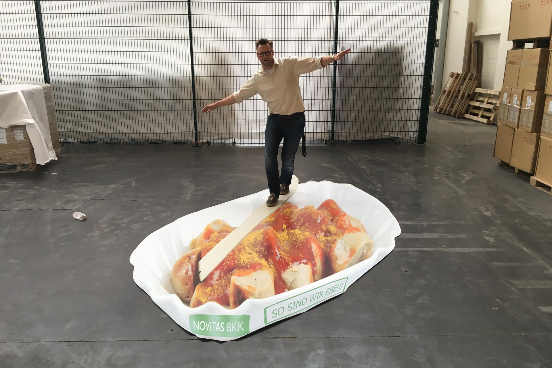 Foto: Marcel Rüffin balanciert mit ausgebreiteten Armen auf einer überdimensionalen Currywurst-Schale mit Werbeaufdruck „Novitas BKK – So sind wir eben!“.