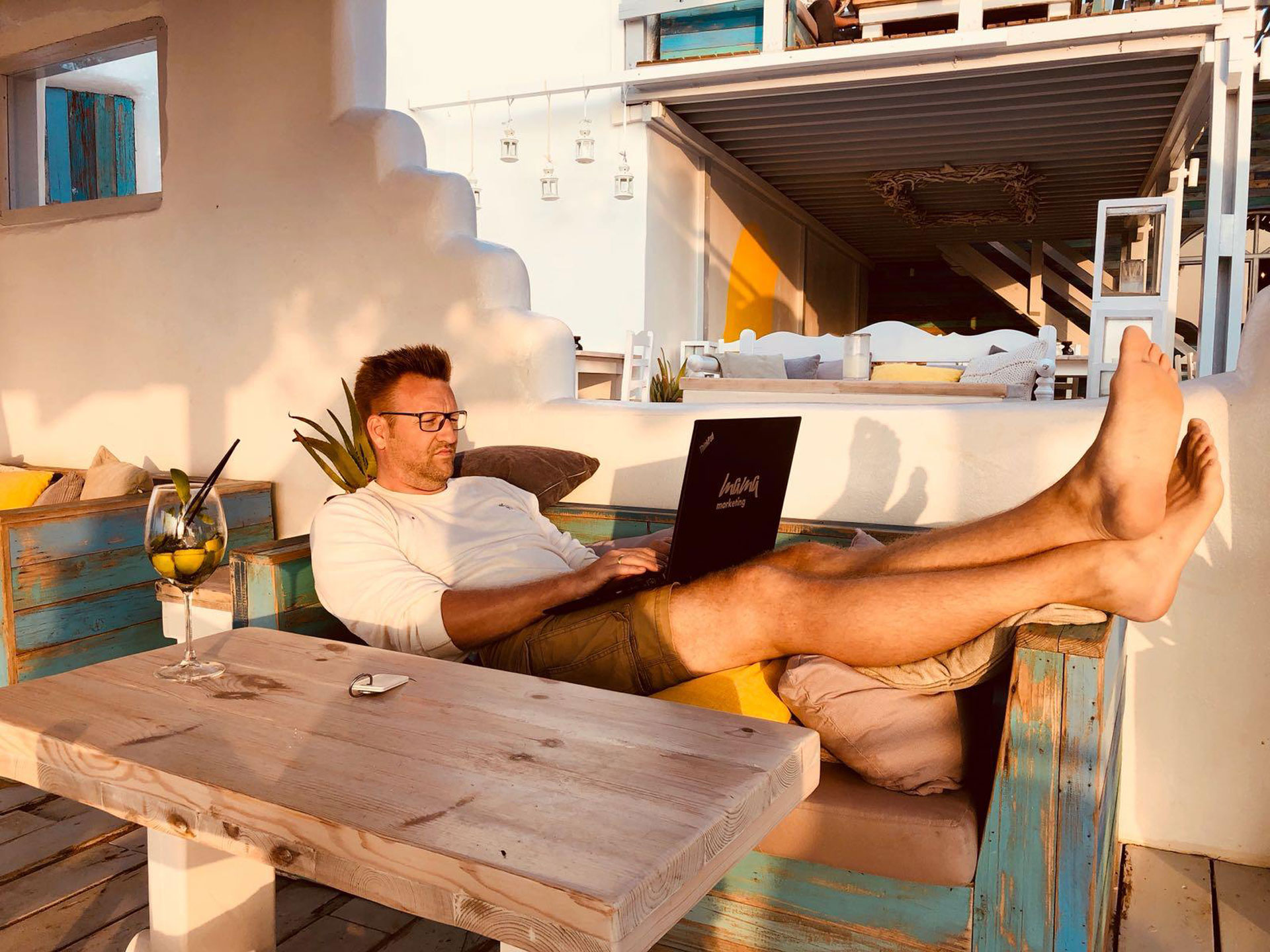 Foto: Marcel Rüffin arbeitet entspannt mit Laptop auf einer rustikalen Sitzbank in mediterranem Ambiente, die Füße hochgelegt, neben ihm ein Drink mit Limetten und Minze.