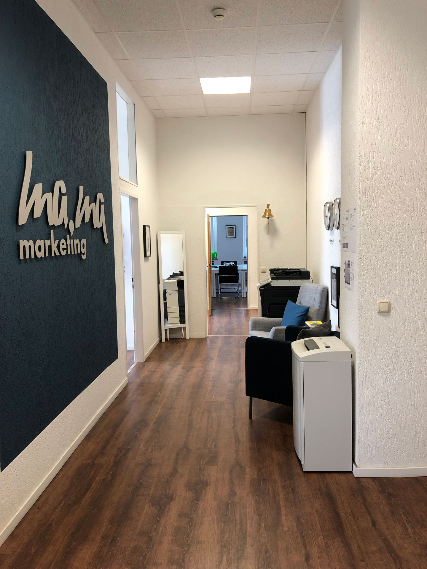 Foto: Moderner Büroflur mit Holzfußboden, Loungeecke und großem „mama marketing“-Logo an der Wand; durch die offene Tür ist ein weiterer Büroraum sichtbar.