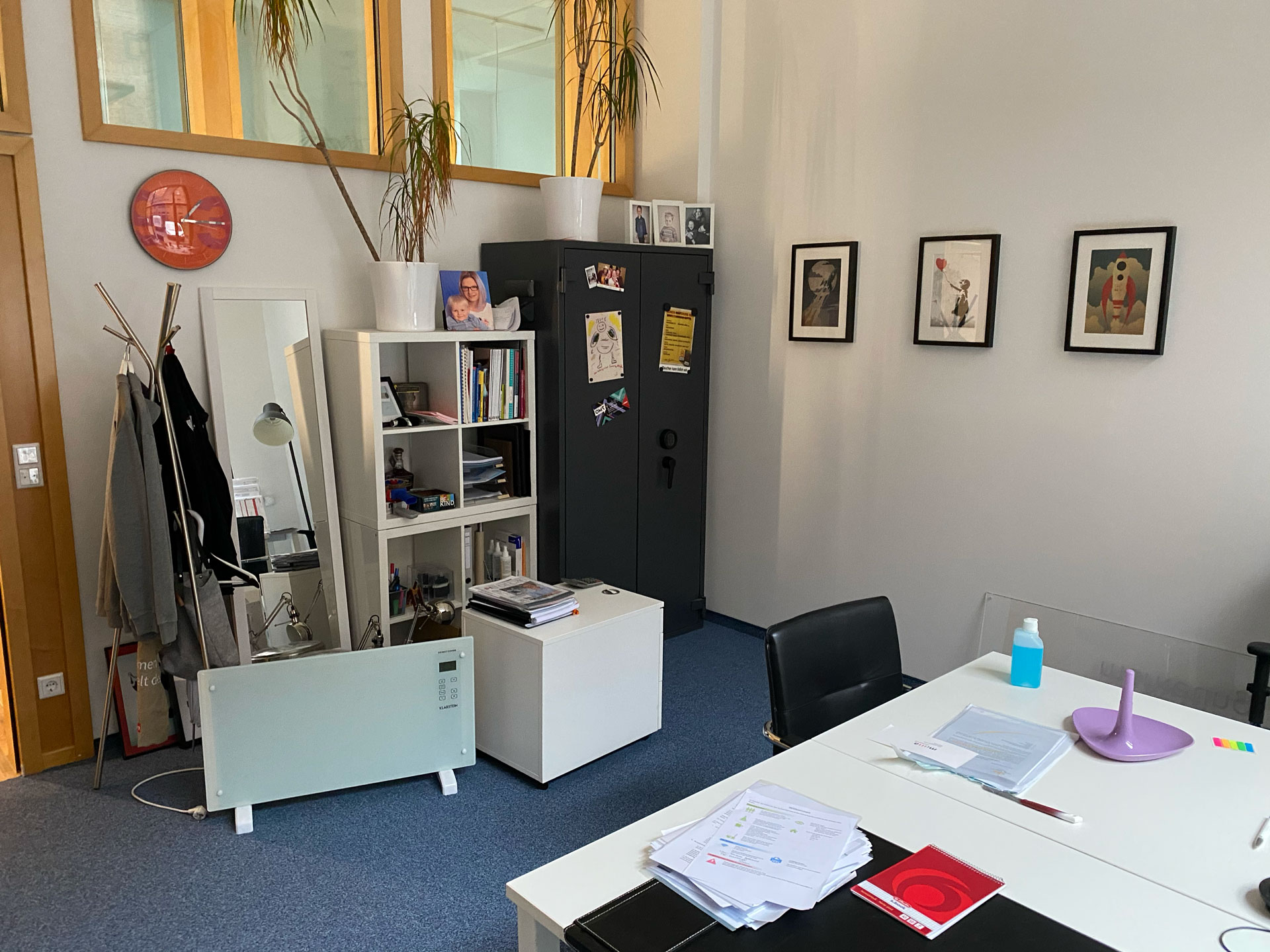 Foto: Individuell eingerichtetes Büro mit Pflanzen, Wandbildern, Bücherregal, Safe und Schreibtisch voller Unterlagen.