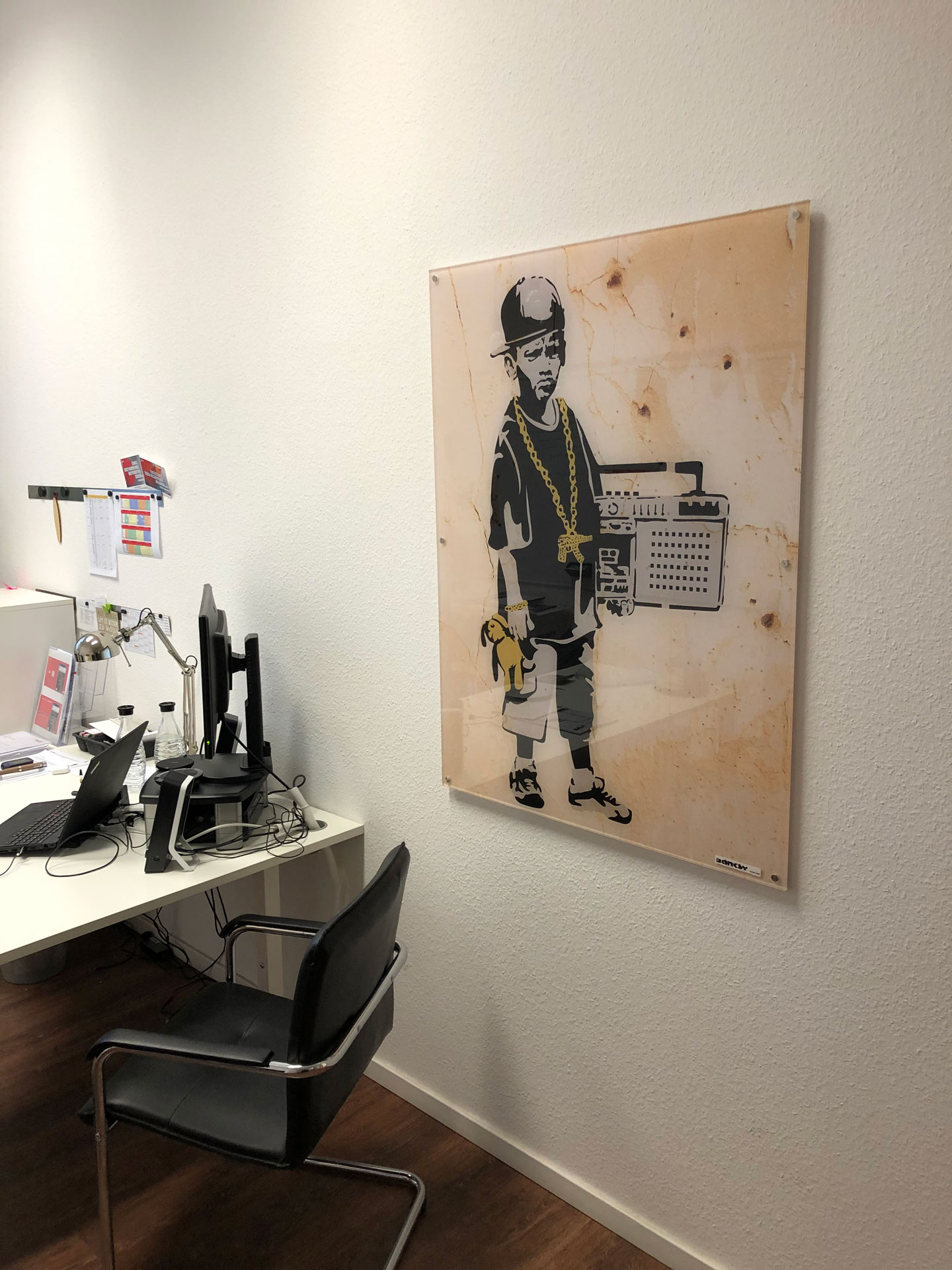 Foto: Arbeitsplatz mit Schreibtisch und einem auffälligen Bild an der Wand, das ein Kind mit Goldkette und Ghettoblaster im Streetart-Stil zeigt.