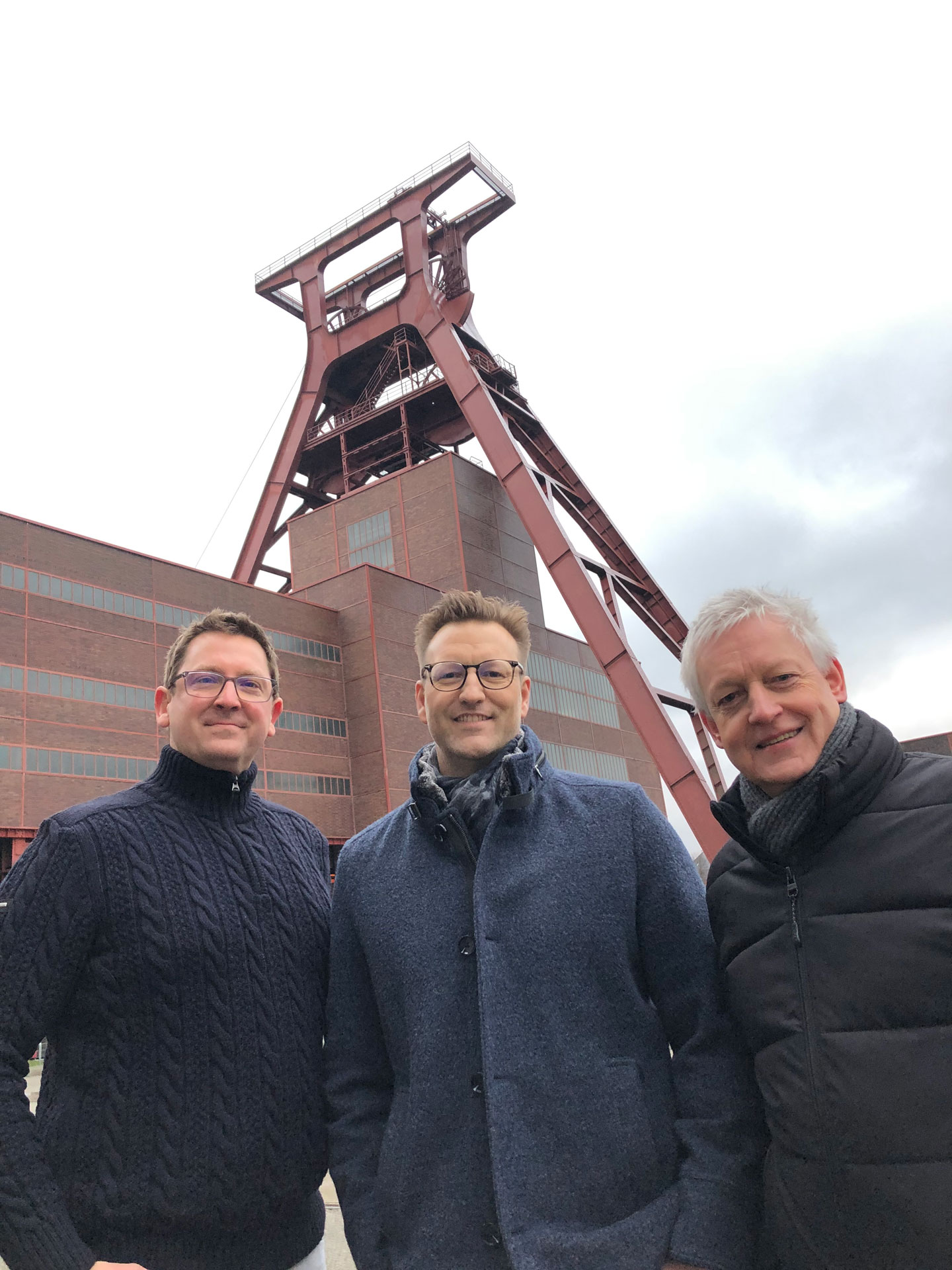 Foto: Drei Männer stehen vor dem Förderturm der Zeche Zollverein in Essen und lächeln in die Kamera.