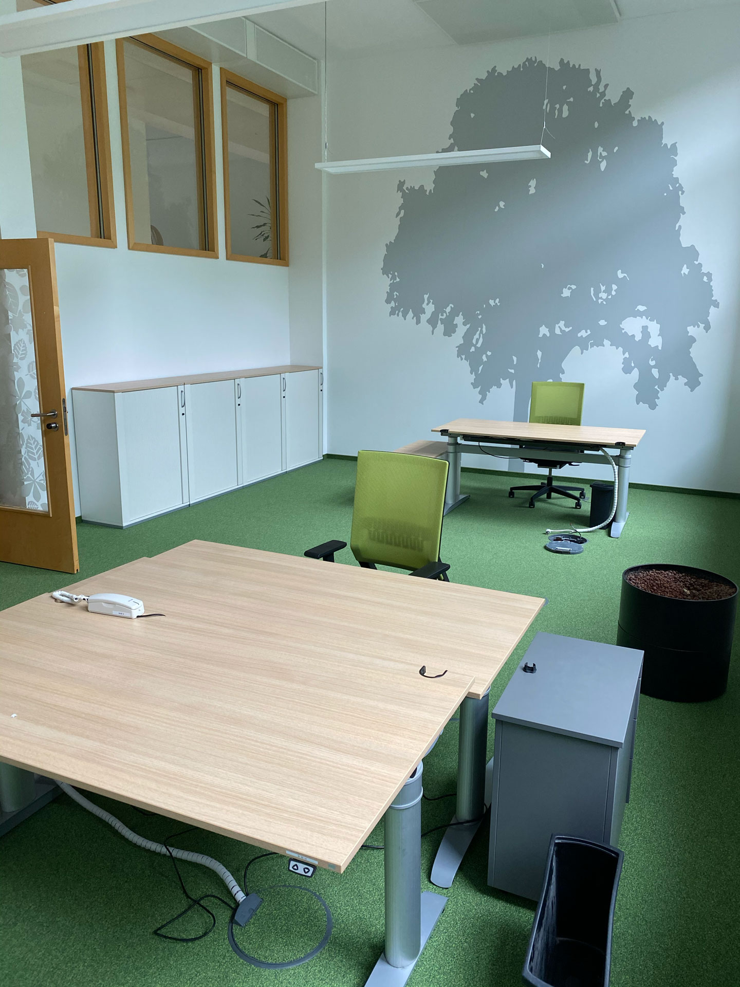 Foto: Modernes, helles Büro mit zwei Schreibtischen, grüner Teppichboden, großen Fenstern und minimalistischer Einrichtung.