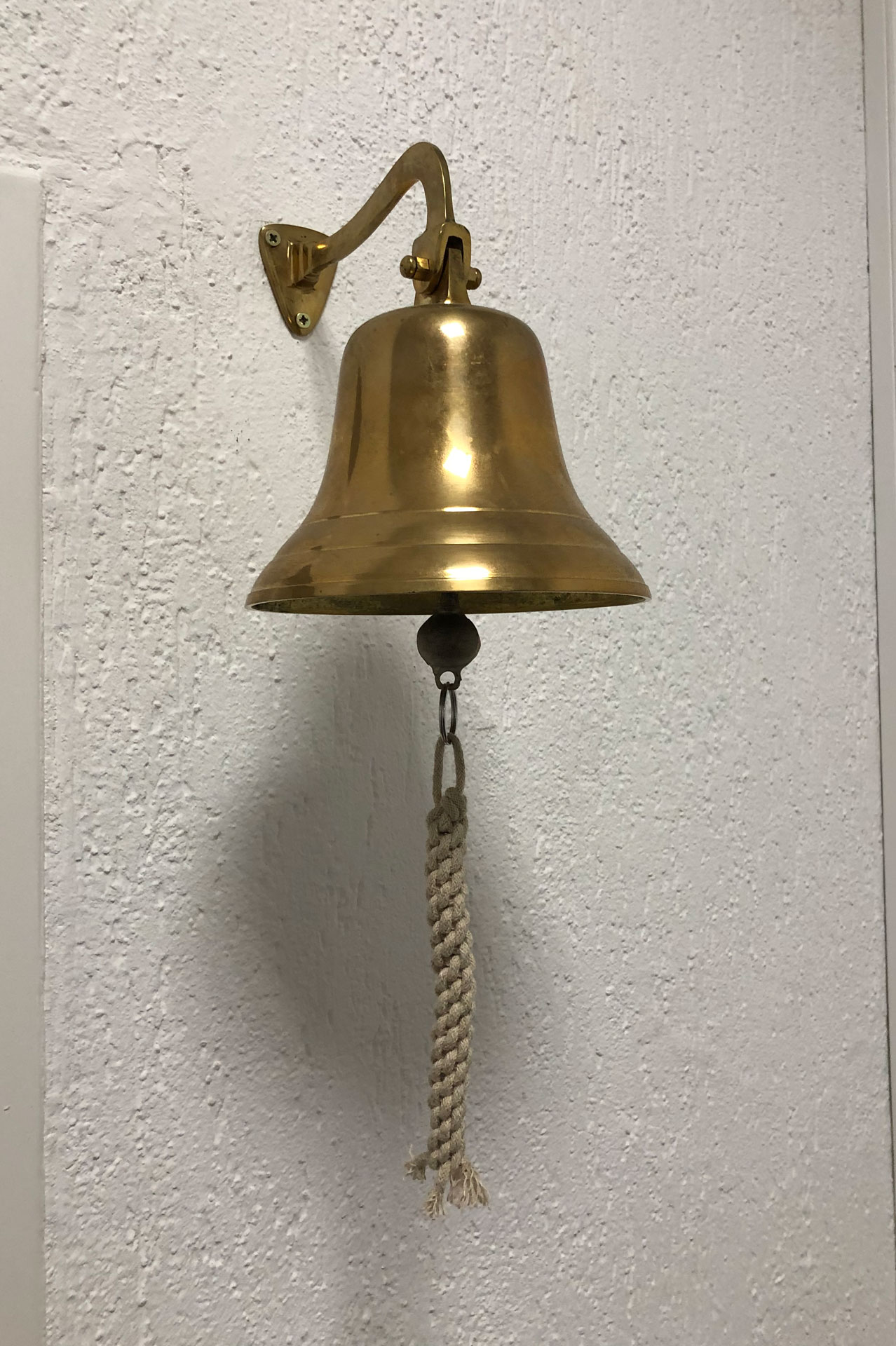 Foto: Goldene Messingglocke mit Kordelzug an einer weißen Wand montiert, erinnert an klassische Schiffs- oder Empfangsglocken.