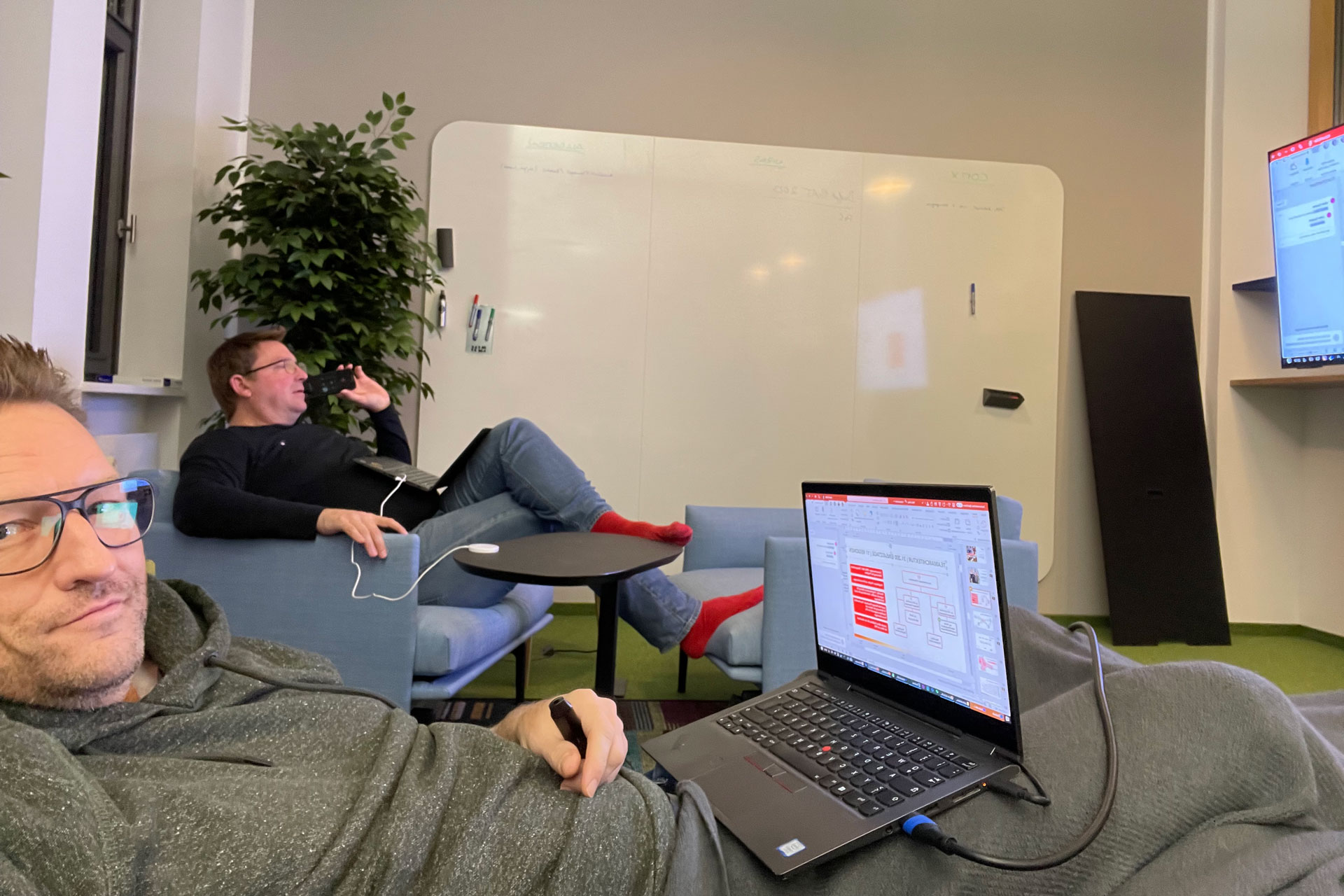 Foto: Marcel Rüffin und Marian Stawski-Hinkelmann arbeiten entspannt mit Laptops auf Sofas, im Hintergrund eine Whiteboard-Wand und große Pflanze.