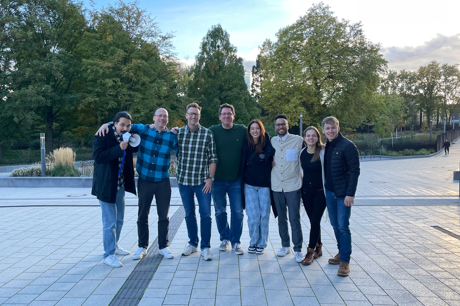 Foto: Gruppe von neun Personen posiert draußen bei Tageslicht auf einer gepflasterten Fläche, alle lächeln.