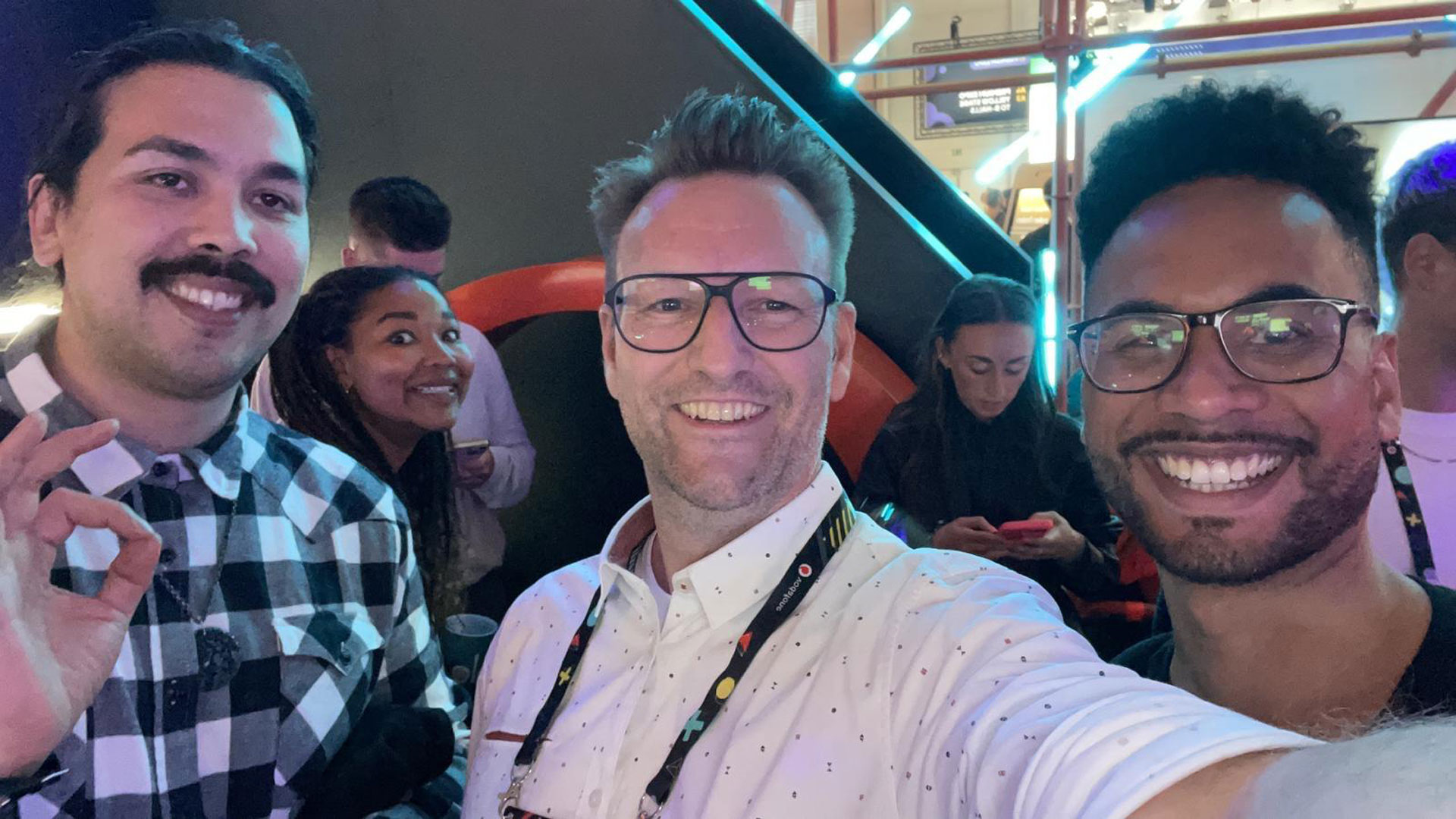 Foto: Drei Männer machen ein Selfie bei einer Veranstaltung mit Neonlicht, im Hintergrund weitere Besucher und bunte Beleuchtung.