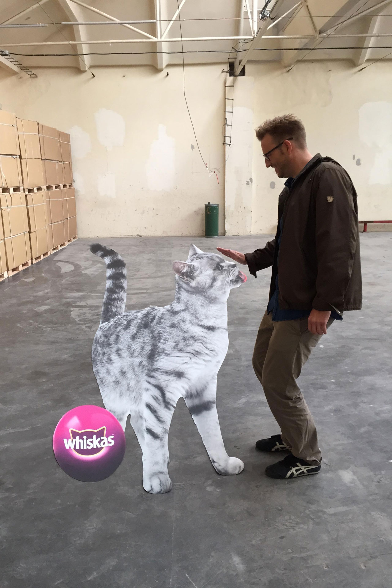Foto: Marcel Rüffin steht in einer Lagerhalle einer lebensgroßen Pappfigur einer Katze mit Whiskas-Logo gegenüber.