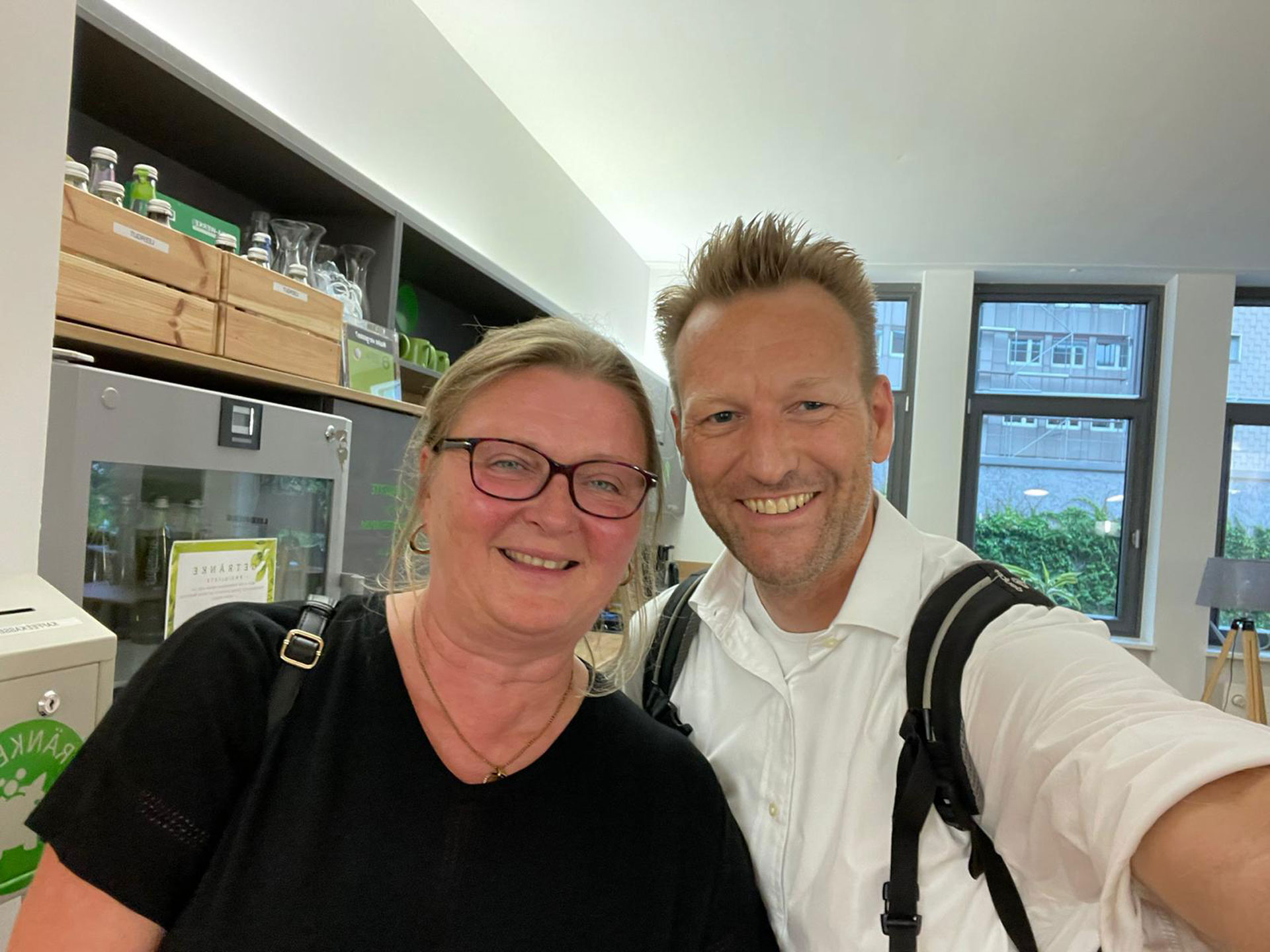 Foto: Frau und Mann machen ein Selfie in einer modern eingerichteten Büroküche, beide lächeln herzlich.