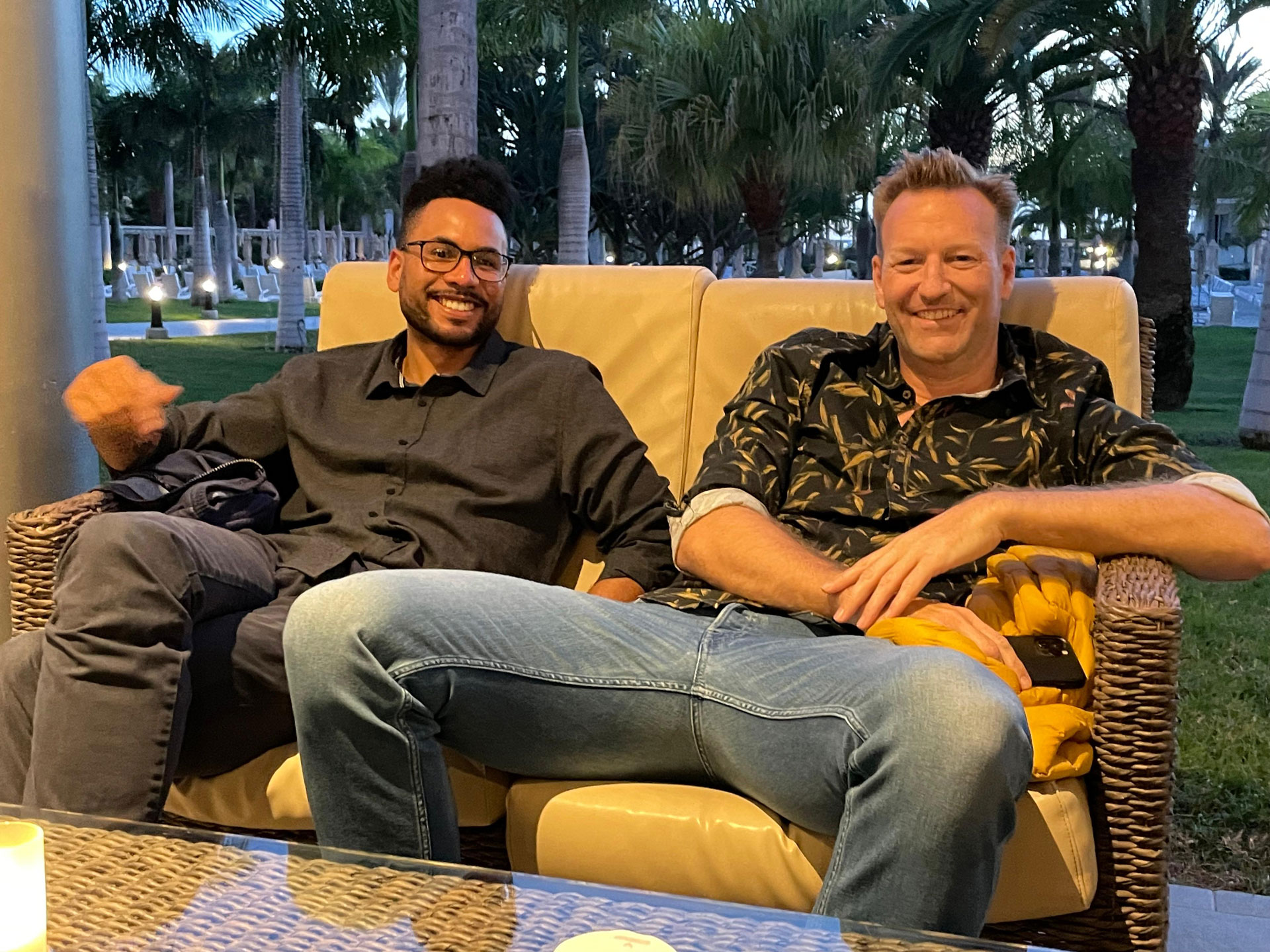 Foto: Zwei Männer sitzen entspannt auf einer Outdoor-Couch unter Palmen, lächeln in die Kamera, eine Kerze brennt auf dem Tisch.