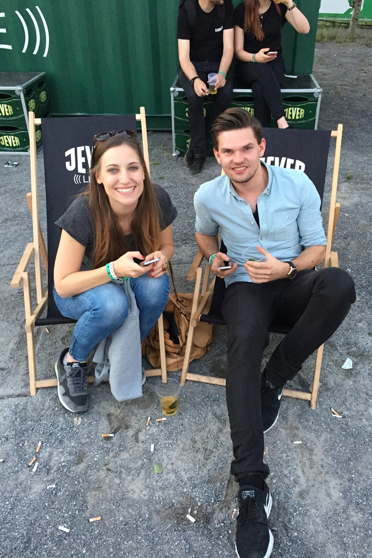 Foto: Junge Frau und junger Mann sitzen lachend in Jever-Liegestühlen bei einem Festival, umgeben von Zigarettenstummeln und Getränken.
