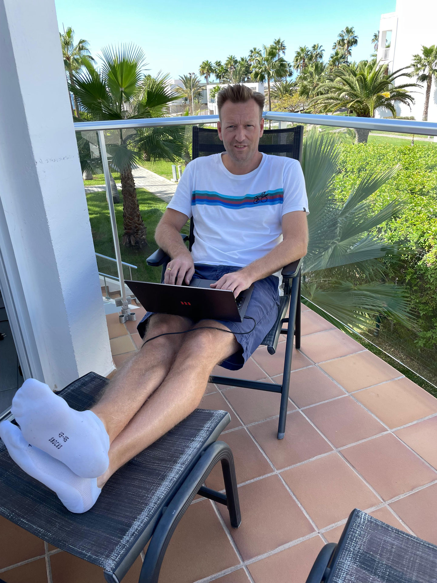 Foto: Marcel Rüffin sitzt entspannt auf einem Balkon mit Laptop auf dem Schoß und Füßen hochgelegt, im Hintergrund Palmen und blauer Himmel