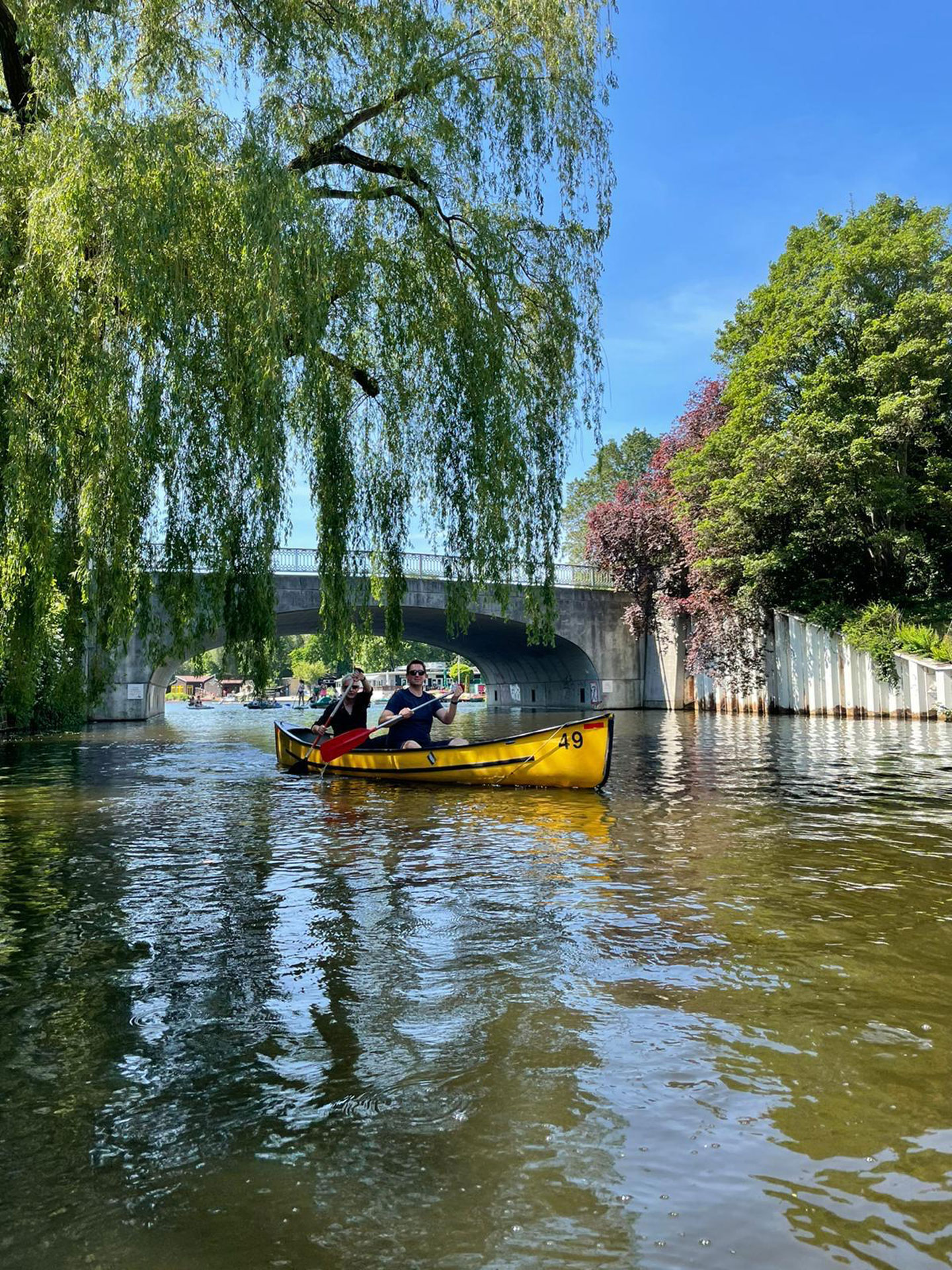 Foto: Zwei Männer paddeln in einem gelben Kanu auf einem sonnigen Fluss unter einer Brücke, umgeben von grünen Bäumen.