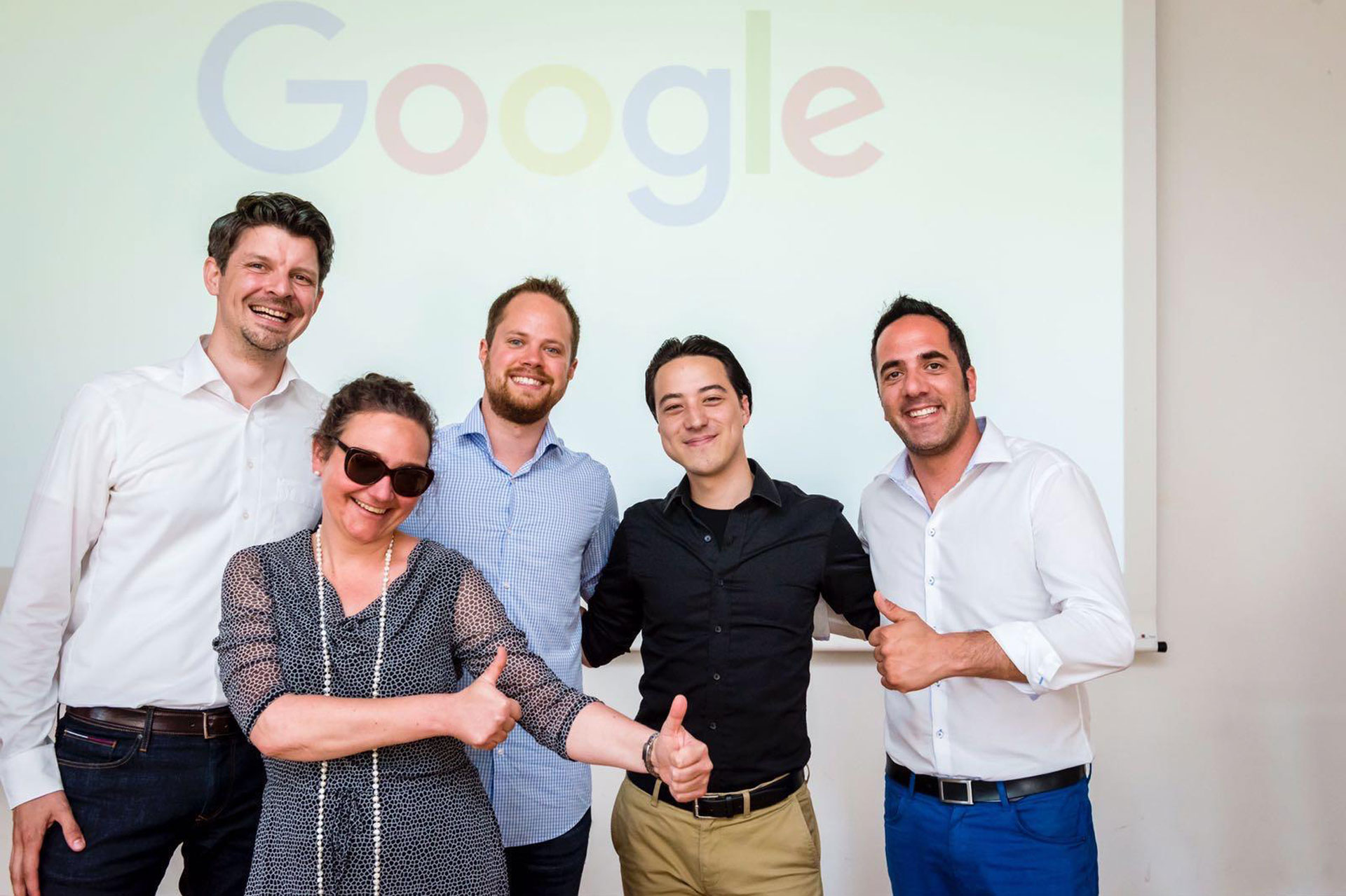 Foto: Fünf Personen posieren fröhlich vor einer Leinwand mit großem Google-Logo, mehrere Daumen hoch.