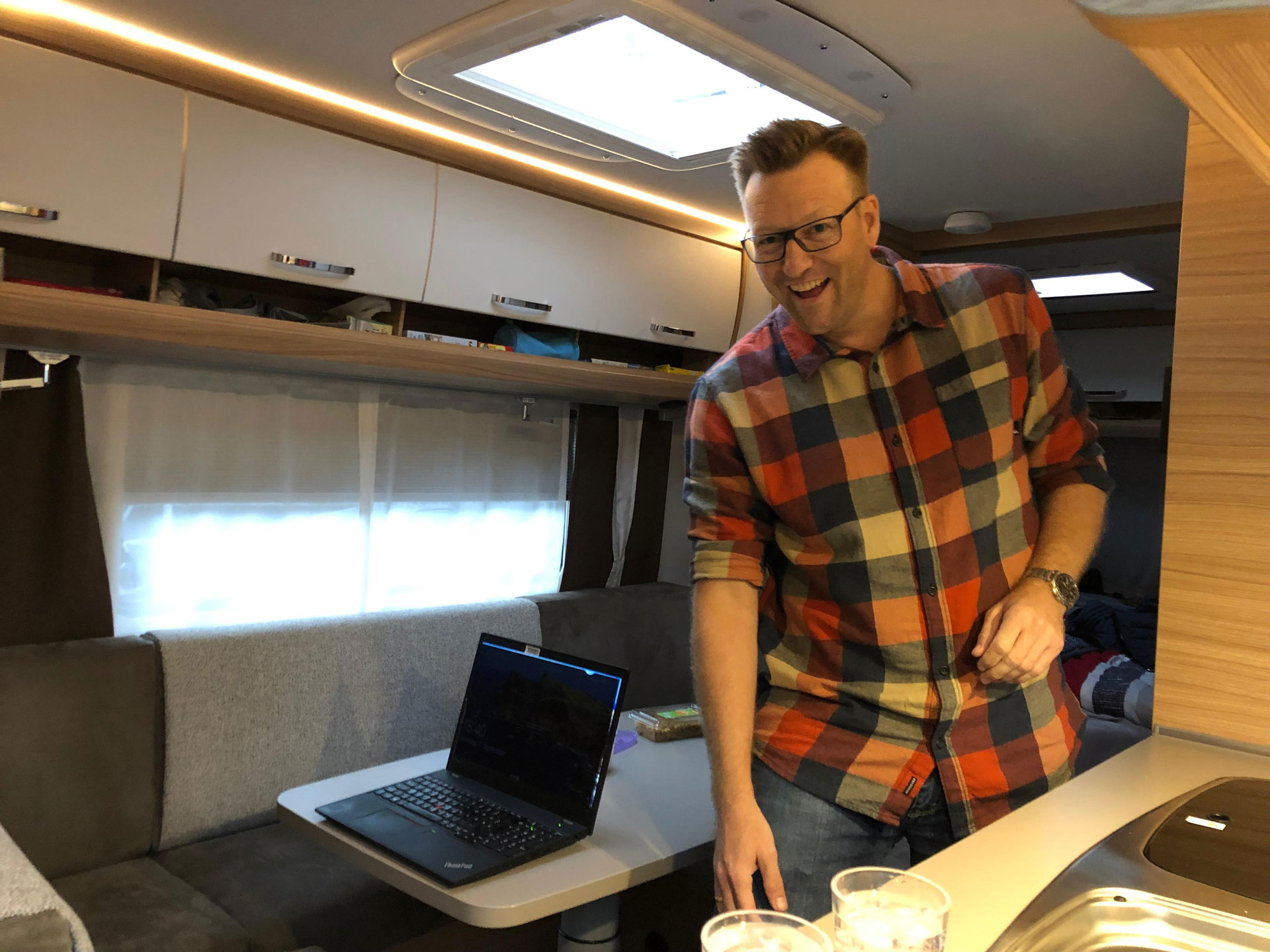 Foto: Marcel Rüffin mit kariertem Hemd steht lachend in einem gemütlichen Wohnmobil, auf dem Tisch vor ihm ein aufgeklappter Laptop.