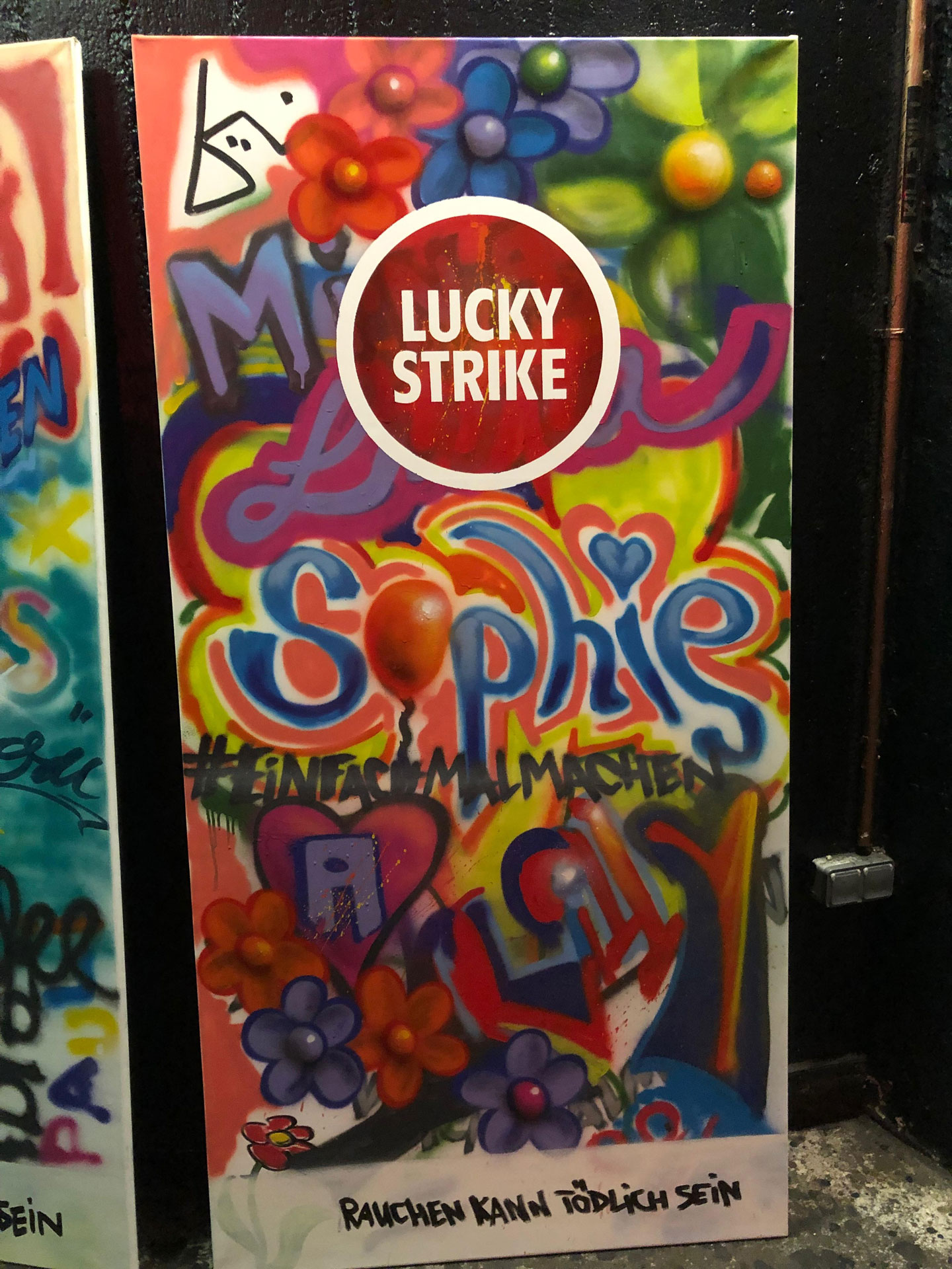 Foto: Buntes Graffiti-Plakat mit „Lucky Strike“-Logo, dem Namen „Sophie“, vielen Blumen und dem Schriftzug „Rauchen kann tödlich sein“.