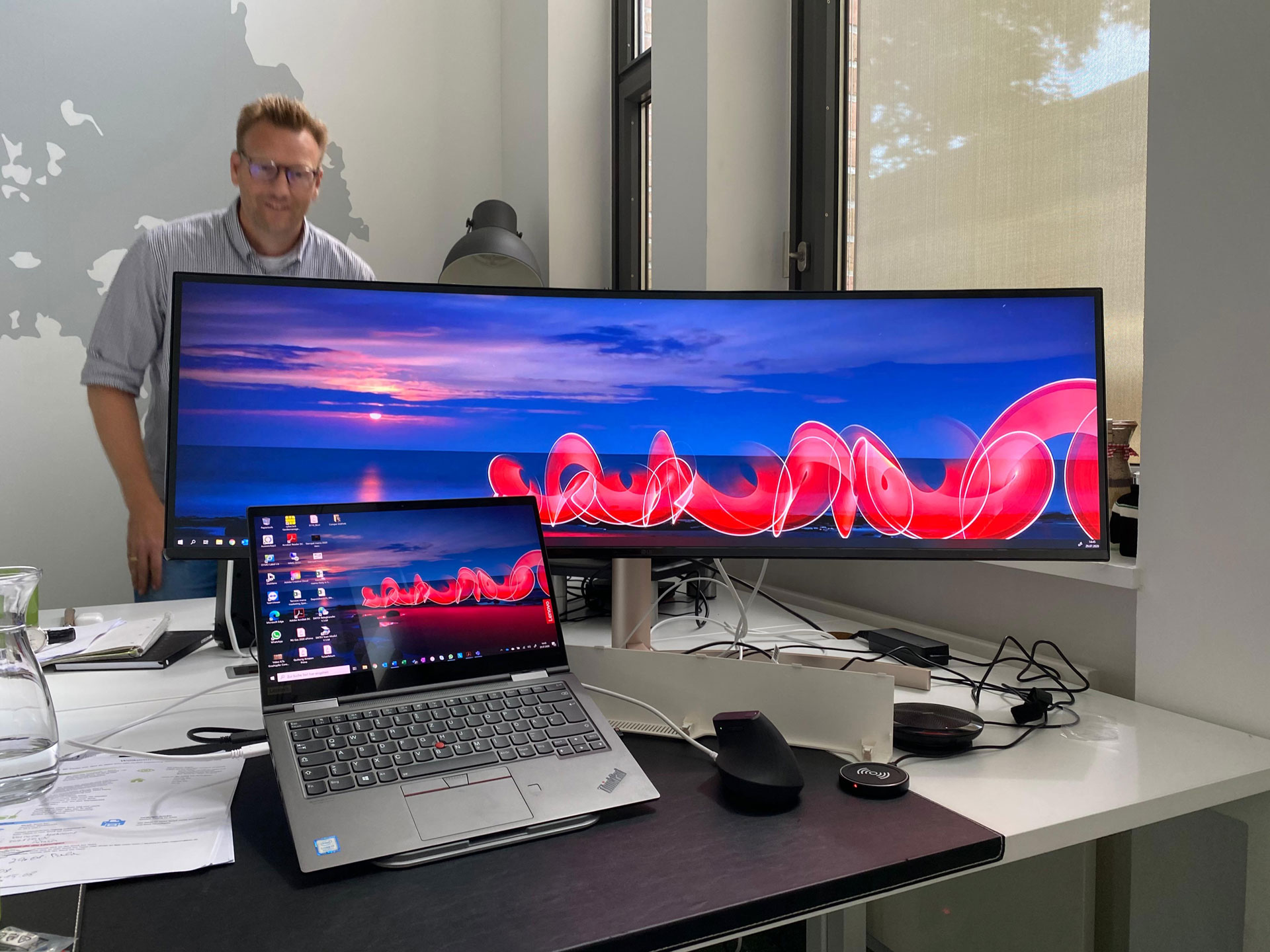 Foto: Marcel Rüffin steht in einem modernen Büro hinter einem Schreibtisch mit ultrabreitem Monitor und aufgeklapptem Laptop; auf beiden Bildschirmen ist ein Sonnenuntergang mit rotem Lichtmuster zu sehen.