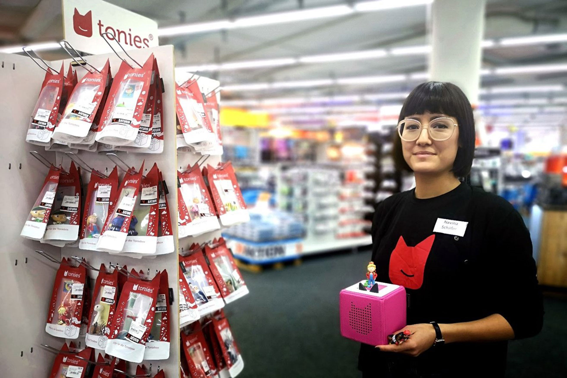 Foto: Promoterin mit Namensschild „Navina“ präsentiert eine pinke Toniebox in einem Elektronikmarkt vor einem Regal mit Tonie-Figuren.