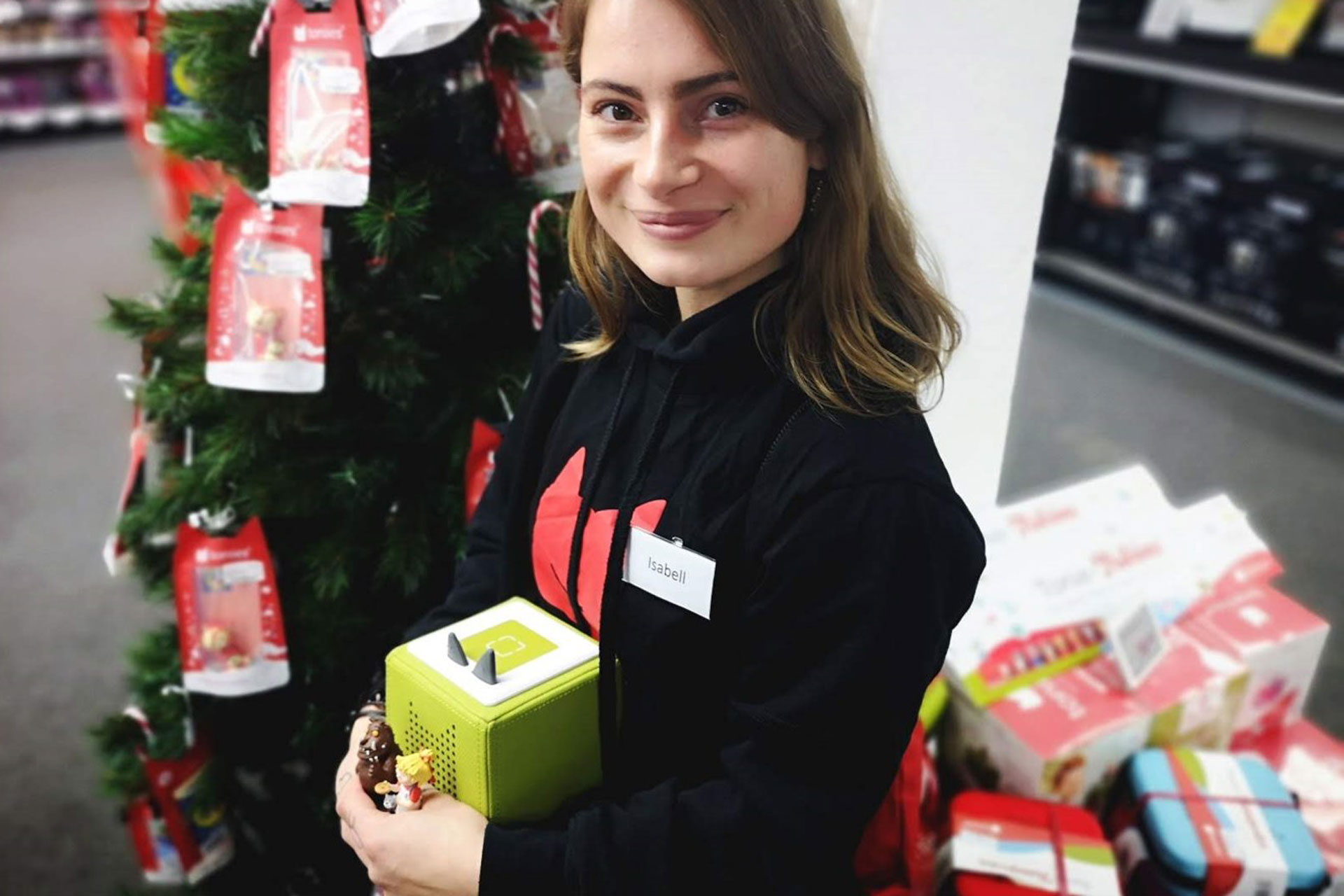 Foto: Promoterin mit Namensschild „Isabell“ hält eine grüne Toniebox in einem weihnachtlich dekorierten Elektronikmarkt.
