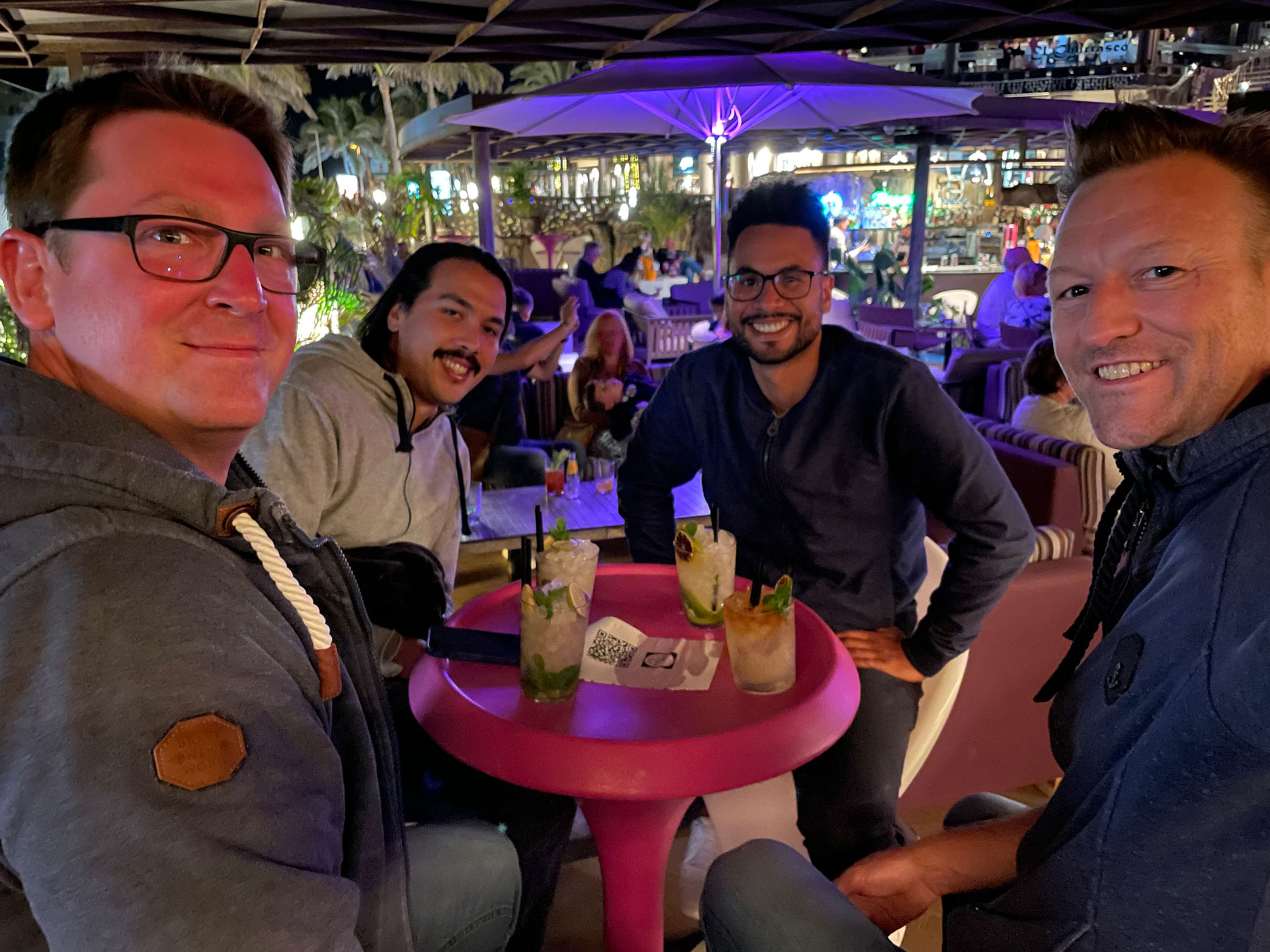 Foto: Vier Männer sitzen bei Nacht an einem runden Tisch in einer belebten Outdoor-Bar, vor ihnen bunte Cocktails, die Stimmung ist entspannt und fröhlich.