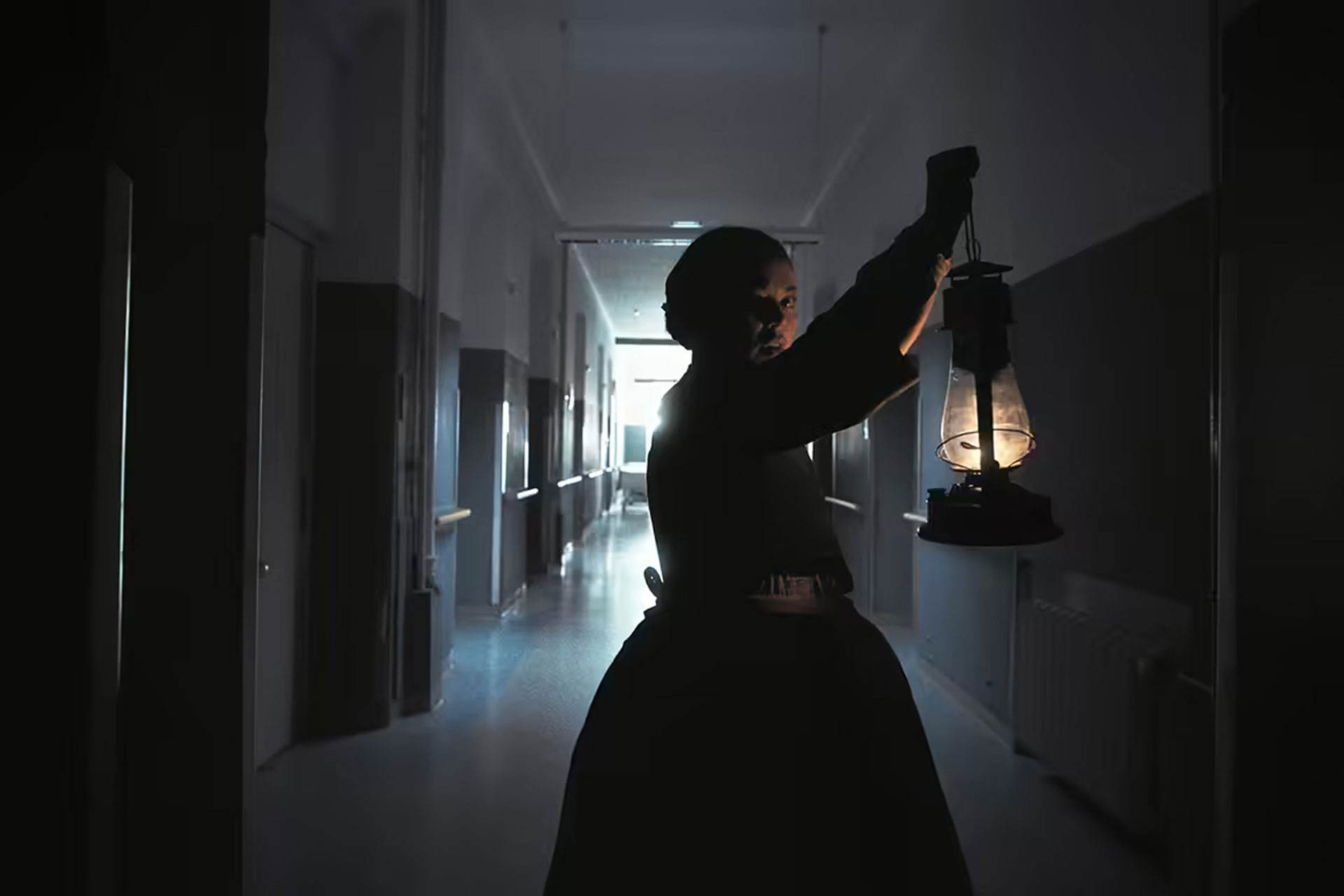 Kachel_DBfK_Film Kachelbild: Eine historische Szene im Stil eines Films – eine Frau im langen Kleid hält eine Petroleumlampe in einem dunklen Flur, das Licht fällt dramatisch auf ihr Gesicht. Ideal zur Darstellung von Themen wie Pflegegeschichte, Mut oder Nachtarbeit.